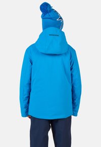 Světlá modrá zateplená bunda s kapucí, s hladkou texturou. K ní patří modrý čepice s bambulí a bílým designem blesku.