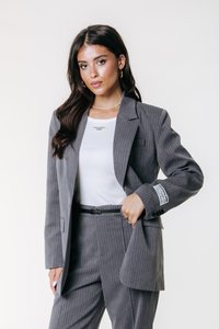Grå stribede blazer båret over en hvid top, med en enkelt brystlomme og knaplukninger ved ærmerne, sammen med slim-fit bukser og et sort bælte.