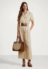 Femme portant une combinaison beige avec une large ceinture, des sandales compensées marron et tenant un sac fourre-tout en osier marron, se tenant devant un fond blanc.