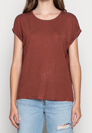 Mujer que lleva una camiseta sencilla de color óxido con mangas cortas y vaqueros rotos azul claro, de pie frente a un fondo liso.