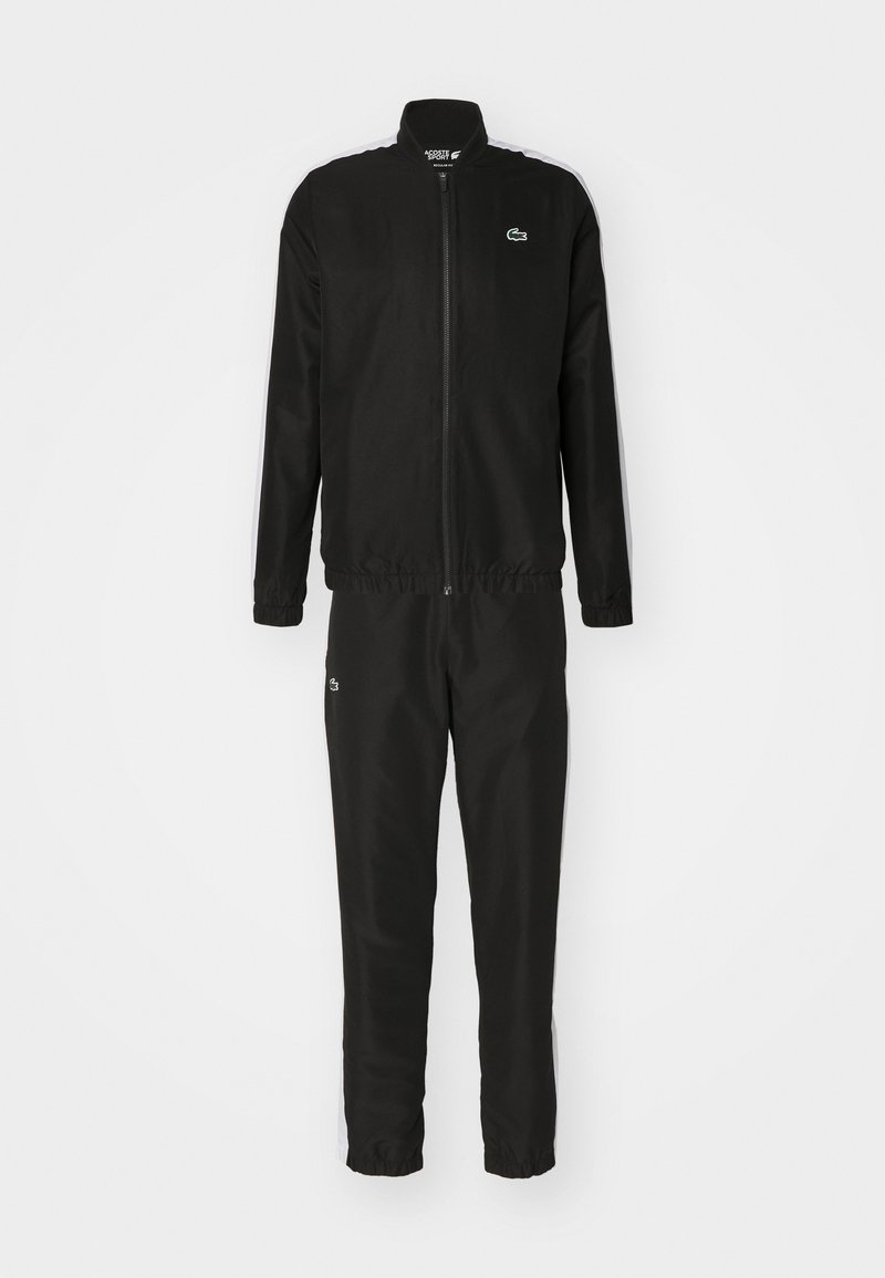 Tuta nera con giacca zip e pantaloni con vita elasticizzata. Presenta un piccolo logo verde e strisce bianche sui lati. Texture liscia.