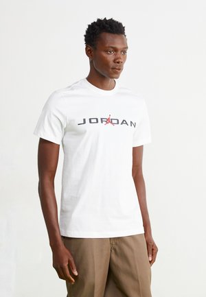 T-shirt en coton blanc avec un logo "JORDAN" noir et rouge accompagné d'une icône. Manches courtes et encolure ronde, texture lisse, coupe décontractée.