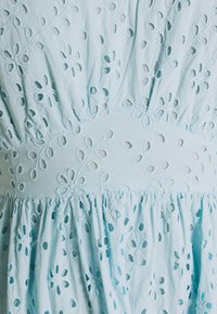 Světle modré vyšívané šaty s květinovými výřezy, staženým pasem a volánkovým lemem. Vyrobeno z měkké, strukturované látky.