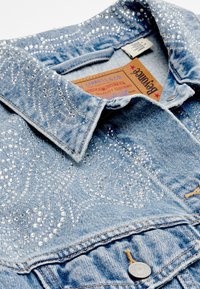 Heled sinine denim jakk, millel on säravad rhinestone kaunistused keerise mustris, klassikaline krae ja metallist nööbikinnitus. Sildid on nähtavad.