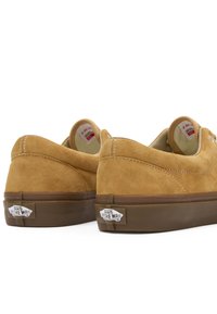 Baskets en daim beige à profil bas, semelle en caoutchouc marron, accents de coutures blanches, et logo argenté "Vans Off The Wall" sur le talon.