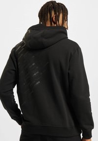 Sudadera negra con un respaldo de patrón texturizado, que presenta logotipos en relieve. Tiene una forma clásica y una capucha con cordón.