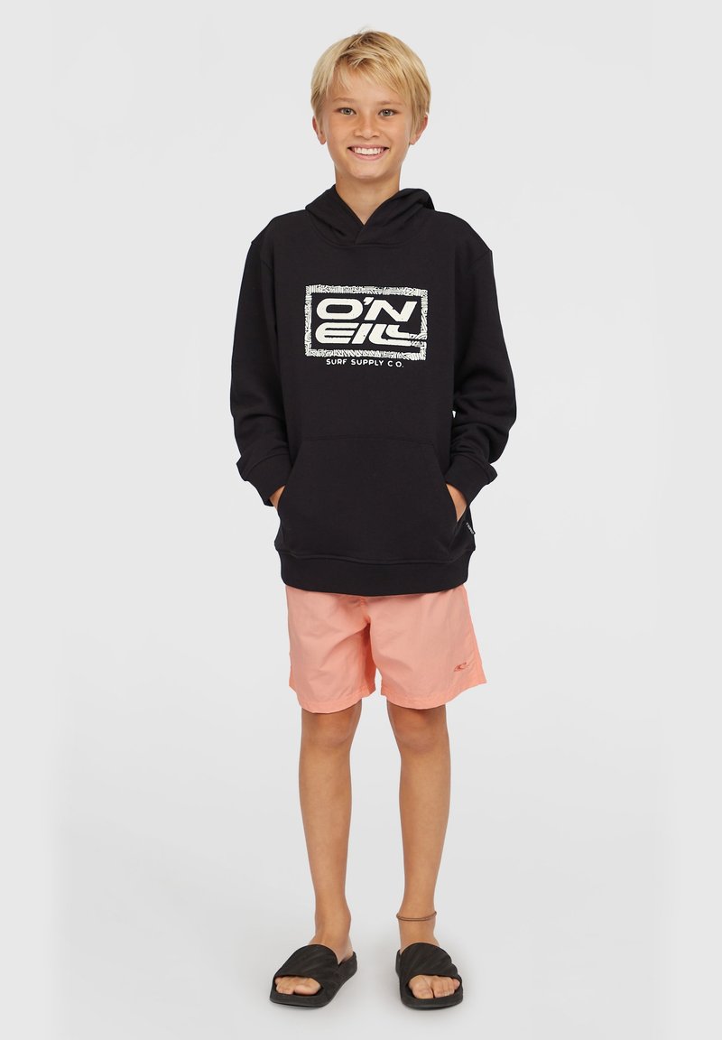 Sudadera negra con capucha y estampado gráfico blanco, combinada con pantalones cortos rosa claro. El sujeto lleva sandalias negras tipo slide.