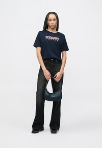 Mornarsko modra bombažna majica s logotipom "tommy jeans", kombinirana s črnimi razširjenimi kavbojkami. Drži mornarsko modro usnjeno torbico z reliefnim logotipom in srebrnimi kovinskimi dodatki.