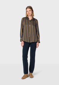 Camicia con bottoni a righe verticali blu navy e senape, maniche lunghe e colletto, abbinata a pantaloni scuri e sneakers color cuoio.