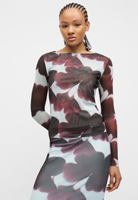 Νέα γυναίκα με πλεξούδες, που φοράει εφαρμοστή μπλούζα μακριού μανικιού και φούστα με μεγάλα αφηρημένα μοτίβα λουλουδιών σε μωβ και γκρι αποχρώσεις.