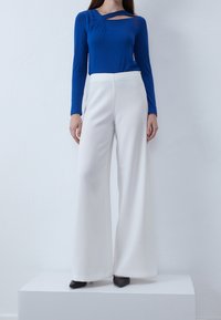 Top blu lungo con maniche e un dettaglio di ritaglio unico abbinato a pantaloni bianchi a gamba larga. Entrambi gli indumenti sono lisci, aderenti e casual. Tacchi neri completano il look.