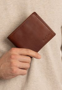Hand holding a brown leather bifold wallet with visible stitching and embossed "Gusti Leder studio" logo on beige fabric background.