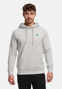 Jonge man met kort krullend haar, gekleed in een lichtgrijze hoodie met voorkantzak en groen logo, staand tegen een witte achtergrond.