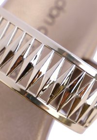 Correa metálica dorada con recortes triangulares, que cuenta con una plantilla suave en color beige. El diseño enfatiza ángulos agudos para una estética audaz.