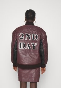 Burgundská kožená bomber bunda s prešívanou textúrou a čiernymi koženými rukávmi, s nápisom "2ND DAY" v tučnom bielom písmene na chrbte.