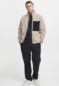Beige fleece jacket with a black patch pocket, zip front, and high collar, paired with black pants and white sneakers. Knit beanie.