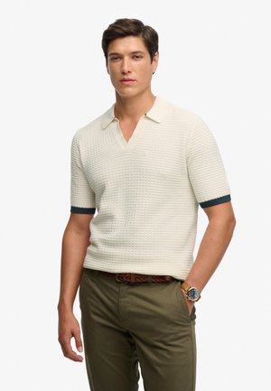 Camiseta tipo polo de manga corta en color crema con patrón texturizado, escote en V, puños en contraste de color verde oscuro, combinada con pantalones oliva y cinturón marrón.