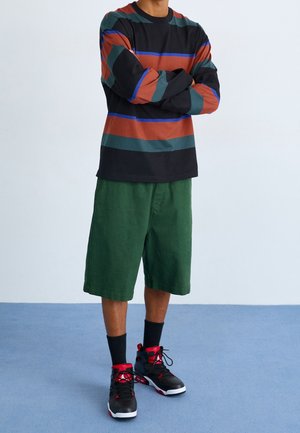 Långärmad randig skjorta i svart, rost, grön och blå. Gröna shorts och svarta sneakers med röda accenter. Enkla bomullsmaterial.