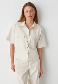 Camicia a maniche corte color crema con due tasche frontali e bottoni in argento. Il tessuto sembra leggero e ha una texture liscia.