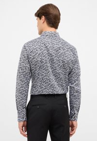 Man met zijn rug naar de camera, draagt een longsleeve shirt met zwart-wit abstract patroon en zwarte broek op een effen achtergrond.