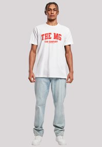 Weißes Baumwoll-T-Shirt mit rotem Drucktext "THE MG CAR COMPANY" auf der Vorderseite, kombiniert mit hellblauen Jeans und weißen Turnschuhen.