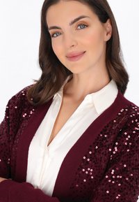 Bordowy kardigan z rękawami ozdobionymi cekinami, zestawiony z białą koszulą z kołnierzykiem. Gładki materiał o luźnym kroju.