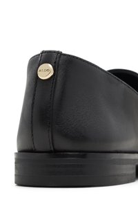 Talons de chaussure habillée en cuir noir avec couture visible et petit symbole rond doré gravé avec "ALDO" sur fond blanc.