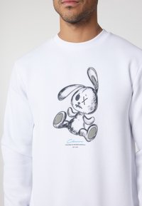 Uomo che indossa una felpa bianca con una grafica di un coniglietto di peluche logoro grigio e nero e un piccolo testo blu sotto nella parte anteriore.