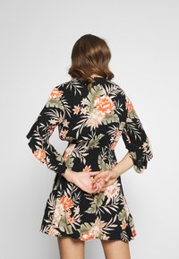 Donna con capelli castani ondulati che indossa un vestito floreale nero, in piedi con le mani incrociate dietro la schiena su uno sfondo semplice.