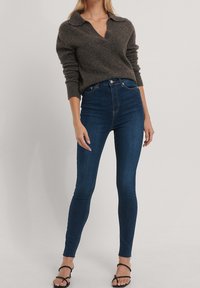 Tmavě šedý pletený svetr s límcem, zkombinovaný s modrými úzkými džíny s vysokým pasem. Modelka má na sobě černé sandály na vysokém podpatku.