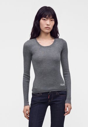 Kvinde med langt sort hår iført en tætsiddende grå ribstrikket langærmet sweater og mørkeblå jeans stående mod en hvid baggrund.