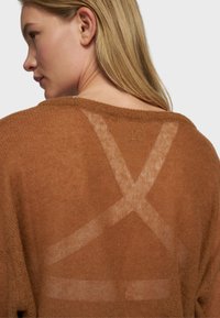 Pull en maille brun avec un dos transparent orné d'un motif en X et le numéro "10" brodé en fil doré, créant un contraste visuel texturé.