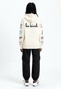 Kremowa bluza z kapturem z napisem "bądź miły" na plecach; kolorowe grafiki na rękawach. W zestawie z czarnymi spodniami bojówkami i białymi tenisówkami.