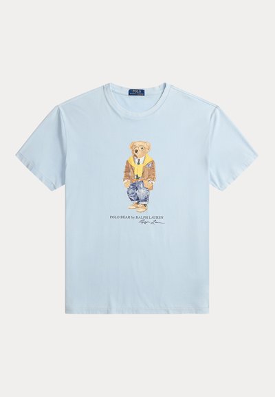 T-shirt azul claro com um urso impresso a usar uma jaqueta castanha, um suéter amarelo, jeans azuis e sapatos bejes, com o texto Polo Bear embaixo.