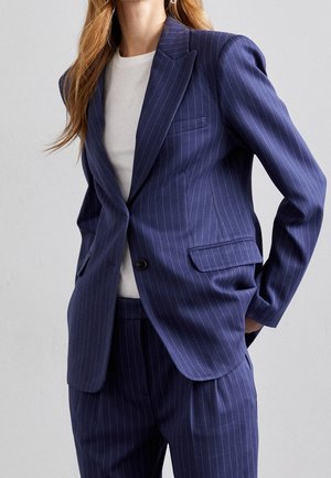 Femme portant un tailleur bleu marine à rayures fines, composé d'une veste à un bouton et d'un pantalon assorti, accompagné d'un t-shirt blanc à col rond.