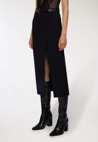 Donna che indossa una gonna midi nera con spacco frontale, vita con cintura e stivali con tacco in pelle nera fino al ginocchio su uno sfondo bianco.
