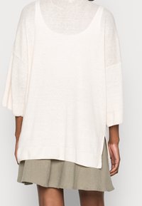 Pull oversize beige clair à manches trois-quarts, poignets côtelés et fentes sur les côtés, porté sur une jupe vert sauge. Texture en maille douce.