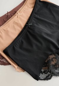 Tre par damunderkläder i svart, nude och brun. Funktionen inkluderar slät tyg, spetskanter och dekorativa rosetter vid midjan.