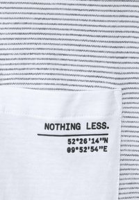 Bílá látka s černými horizontálními pruhy. Bílý nášivka obsahuje text "NIC MÉNĚ." a souřadnice: 52°26'14"N 09°52'54"E.