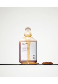 Bottiglia di shampoo ambra trasparente con design arrotondato. Etichettatura chiara su sfondo bianco. Il liquido fuoriesce dalla bottiglia su una superficie.