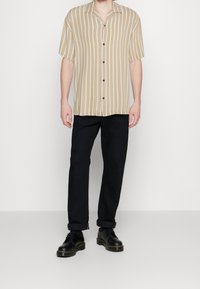 Camicia a maniche corte beige a righe con colletto, davanti con bottoni, abbinata a pantaloni neri e scarpe nere con i lacci.