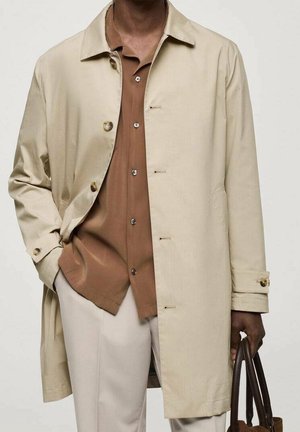 Mężczyzna w beżowym płaszczu trenczu na brązowej, zapinanej na guziki koszuli i jasnych spodniach, trzymający w prawej ręce brązową skórzaną torbę.