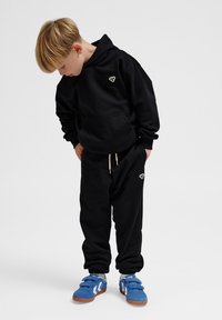 Schwarzes Hoodie- und Jogginganzug-Set mit einer Fronttasche. Mit einem kleinen Logo auf der Brust. Blaue Sneakers mit einem weißen Design und Klettverschlüssen.
