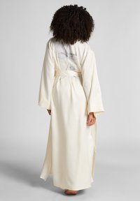 Vrouw met krullend haar, die een crèmekleurige lange satijnen robe met een subtiele bloemenprint op de achterkant draagt, staande op blote voeten op een grijze vloer.