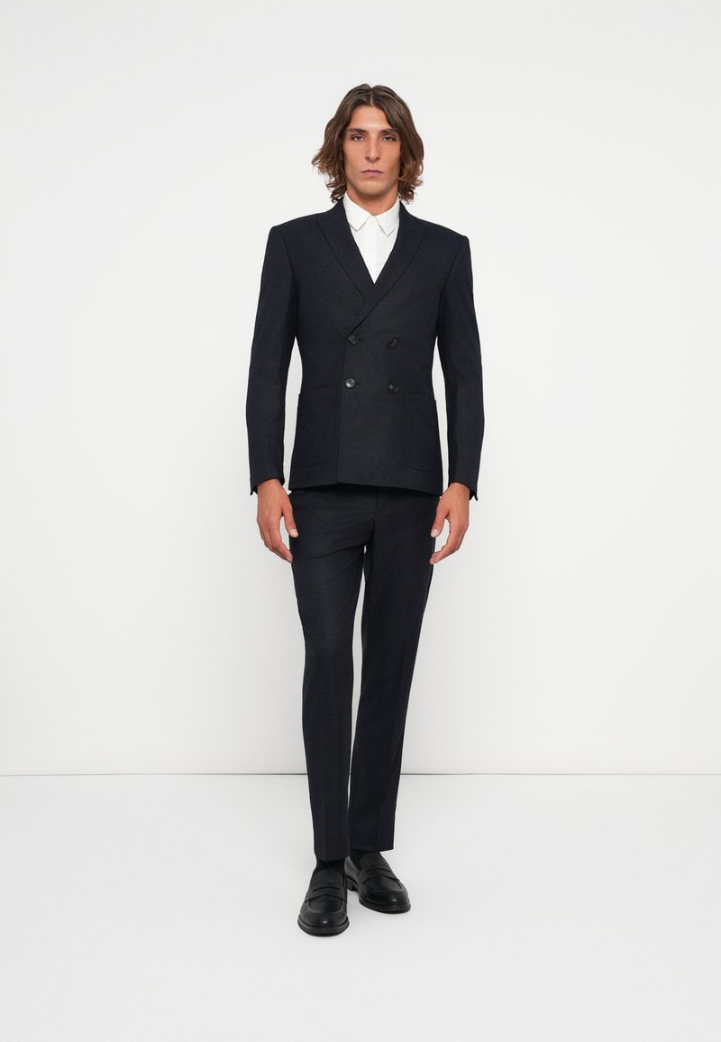 Traje azul marino de doble botonadura con un acabado texturizado, complementado con una camisa blanca. Presenta un corte ajustado y clásicos mocasines negros.