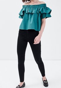 Haut turquoise à épaules dénudées avec un ourlet à volants, fabriqué en tissu léger ; associé à un pantalon noir ajusté et des chaussures noires avec un accent décoratif.