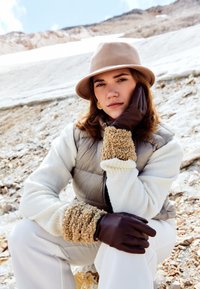 Guantes de cuero marrón con puños de ante texturizados, un suéter de forro polar crema y un chaleco acolchado gris. Un sombrero beige completa el atuendo invernal.