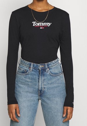 La personne porte un t-shirt noir à manches longues Tommy Jeans New York avec un logo blanc et rouge et un jean taille haute bleu clair avec bouton et fermeture éclair.