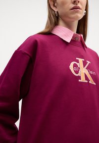 Bordový sveter s vyšívaným logom "Calvin Klein" v oranžovej farbe. Má kontrastný prúžkovaný golier a uvoľnené rukávy.