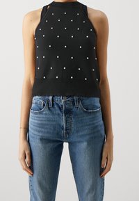 Svart ärmlös topp med pärlprydnader, åtsittande design, ribbad textur och scallopad nederkant, matchad med högmidjade blå denimjeans.